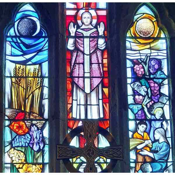 An image of a colourful triptych stained glass window plus celtic cross above the altar in Tintagel Church, aka St Materiana, in Cornwall.