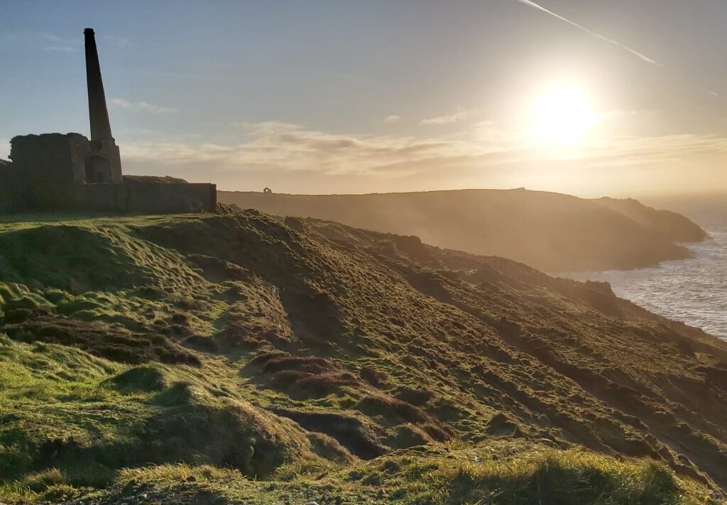 coastal view into the sun featuring grassy hills and a ruined Cornish Engine house